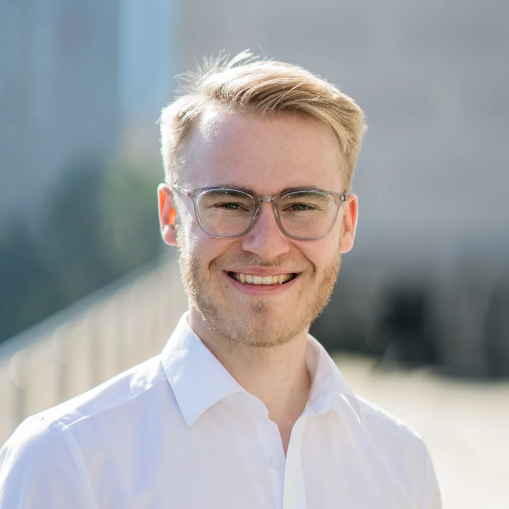 Max Thieme, Geschäftsführer von Carport Frankfurt, lächelt selbstbewusst. Mit blondem Haar, Bart und einer Brille aus transparentem Kunststoff strahlt er Vertrauen und Professionalität aus.